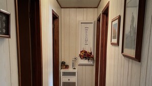 Een gang met houten paneelwanden, een witte deur met een welkomstbord, een klein wit tafeltje met een bloevas en ingelijste foto’s aan de muur.