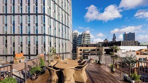 En tagterrasse med et rundt bord og stole, omgivet af plantekasser, med udsigt over en by med moderne bygninger.