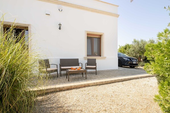 A building with a white exterior, a paved patio area with two chairs and a table, and a car parked nearby. - RELAX SALENTO GALLIPOLI Villa: Terrace, Ocean View, Wi-Fi (Torre Suda)