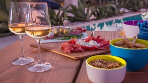 Ein Tisch ist gedeckt mit Weingläsern, einem Holzbrett mit verschiedenen Käse- und Fleischsorten sowie Schalen mit Nüssen und Oliven. Im Hintergrund ist ein Pool und ein Sonnenuntergang zu sehen.