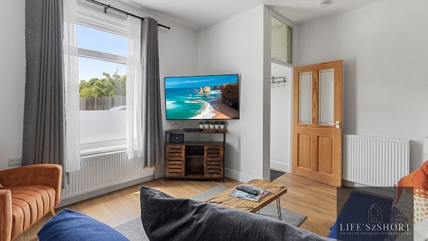 A living room with a television displaying a scenic beach, a wooden cabinet, and a door with a glass panel. - City Gate Garden Lodge (Exeter)