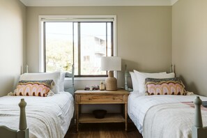 A bedroom with two beds, a nightstand, a lamp, and a window with a view of buildings outside.