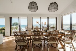 A dining area with a view of the sea, featuring wicker chairs, a wooden table, and hanging pendant lights.