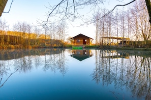 A cabin by a calm lake with a reflection of the trees and cabin. - Arcadia Safari Tent in Private 5 Acre Field (Wrexham)