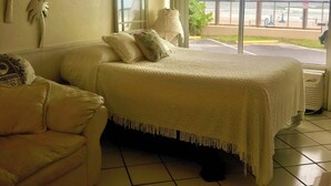 Ein Schlafzimmer mit Bett, Sofa, großem Fenster und Blick auf den Strand sowie Wandbildern mit Palmenmotiven.