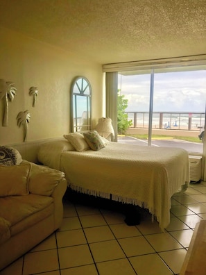 A bedroom with a bed, a sofa, a large window with a view of the beach, and palm tree wall art.