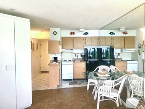 A compact kitchen with white cabinets, a black refrigerator, a stove, and a dining area with white wicker chairs.