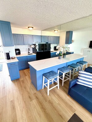 A modern kitchen with blue cabinets, a black refrigerator, and a marble countertop. There are two bar stools and a blue sofa with a striped pillow. - Beach Haven -Ocean View At Symphony Beach Club (Ormond Beach)