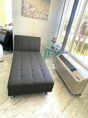 A room with a grey sofa, a small table with a lamp, a radiator, and a window with a view of greenery.