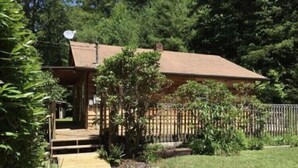 Un chalet rustique doté d’une terrasse en bois, entouré d’une végétation luxuriante et d’une cour clôturée.