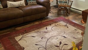 Un salon avec un canapé en cuir marron, des coussins décoratifs à motifs, un tapis rouge et beige, une chaise en bois et des rideaux à carreaux aux fenêtres.