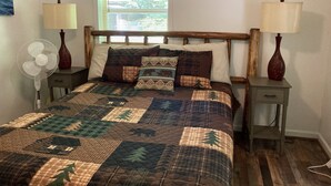 Une chambre à coucher avec une tête de lit en bois, une couvre-lit matelassé, deux tables de chevet avec des lampes, un tableau accroché au mur et une fenêtre avec des stores.