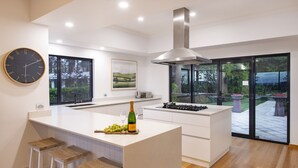 A modern kitchen with a large island, bar stools, a clock, and a view of the outdoors. - LUXURIOUS & INVITING TWO STOREY HOME WITH COMFORTABLY CASUAL COASTAL VIBE (Marybrook)