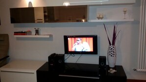 A living room with a flat-screen TV, speakers, a white cabinet, a shelf with books and a vase, and a mirror above the TV. - HEART OF FLORENCE APARTMENT (Firenze)