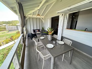 Ein überdachter Essbereich im Freien mit einem Tisch für zwei Personen, umgeben von Stühlen, einer kleinen Couch und einem Blumentopf.
