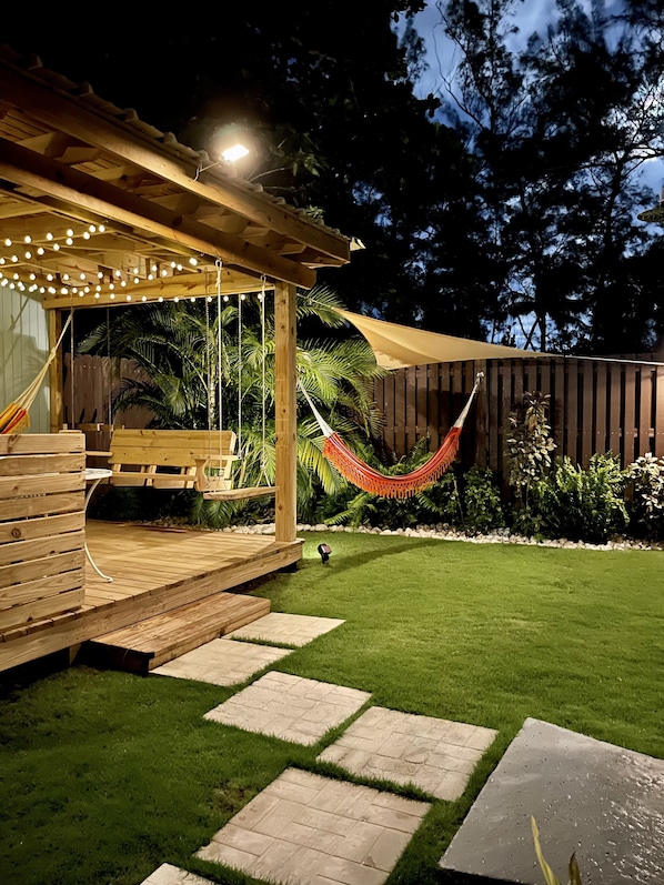 Una terraza de madera con luces colgantes, una hamaca y un espacio de patio con mesa y sillas.