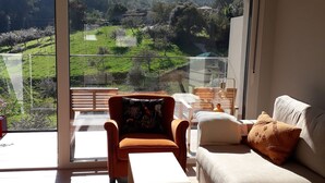 Een moderne woonkamer met een bank, fauteuil en salontafel, met een grote schuifglazen deur die leidt naar een balkon met een prachtig uitzicht op groene velden en bomen.