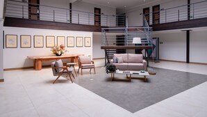 Un vaste espace de vie comprenant un banc en bois, un canapé et une table basse. Un escalier relie les deux niveaux, et des tableaux encadrés ornent le mur.