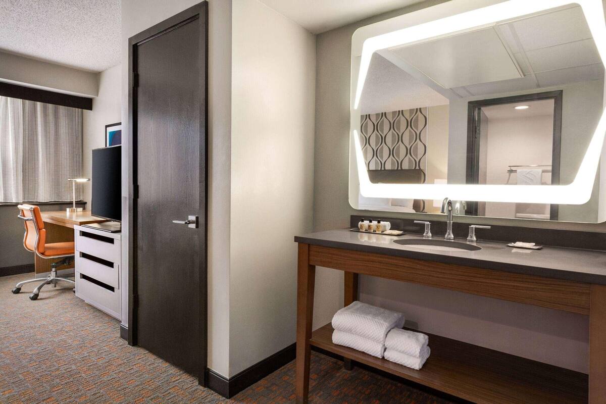 A hotel room with a desk, chair, and a large mirror above a vanity with a sink.