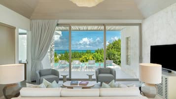 A modern hotel room with a large bed, two armchairs, a coffee table, and a TV. The room features a large window with a view of the ocean and a balcony with outdoor seating.