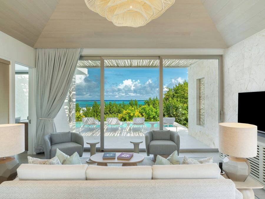 A modern hotel room with a large bed, two armchairs, a coffee table, and a TV. The room features a large window with a view of the ocean and a balcony with outdoor seating.
