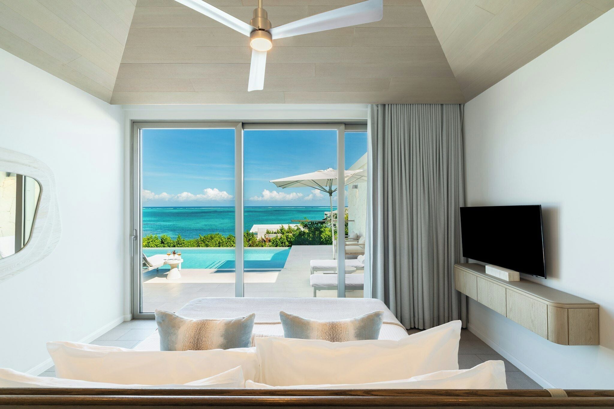 A bedroom with a large window, a ceiling fan, a TV, and a view of the ocean.