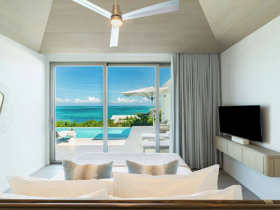 A bedroom with a large window, a ceiling fan, a TV, and a view of the ocean.
