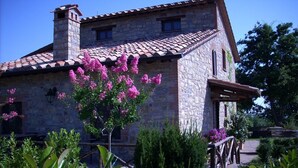 A stone building with a tiled roof, a wooden porch, and a garden with pink flowers.