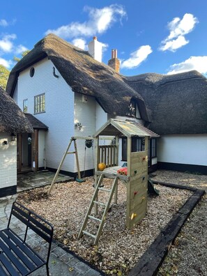 A house with a thatched roof, a wooden play structure, and a bench. - Galtons Cottage Dorset (West Moors)