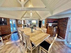 A spacious dining area with a long wooden table and chairs, a brick wall, and a view through a glass door.