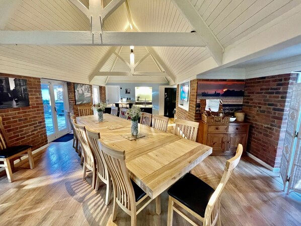 A spacious dining area with a long wooden table and chairs, a brick wall, and a view through a glass door.