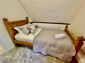 A single bed with a wooden headboard, a grey and white patterned pillow, a white pillow, and a rolled white towel. - Galtons Cottage Dorset (West Moors)