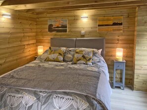 Une chambre dans un chalet en bois, avec un lit, deux lampes fixées au mur et deux tableaux encadrés.