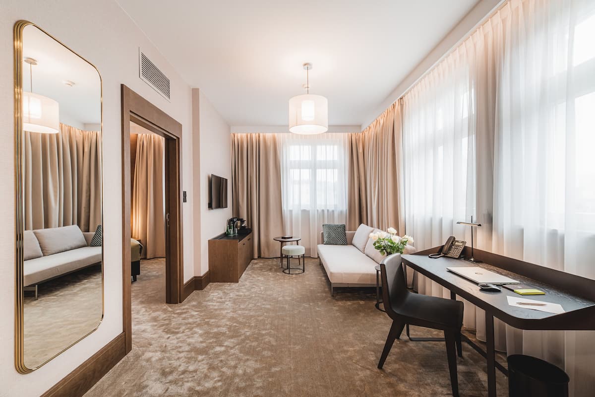 A modern hotel room with a sofa, a desk, a chair, a mirror, and a large window with curtains.