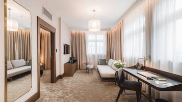 Una habitación de hotel moderna con sofá, escritorio, silla, espejo y un ventanal con cortinas.