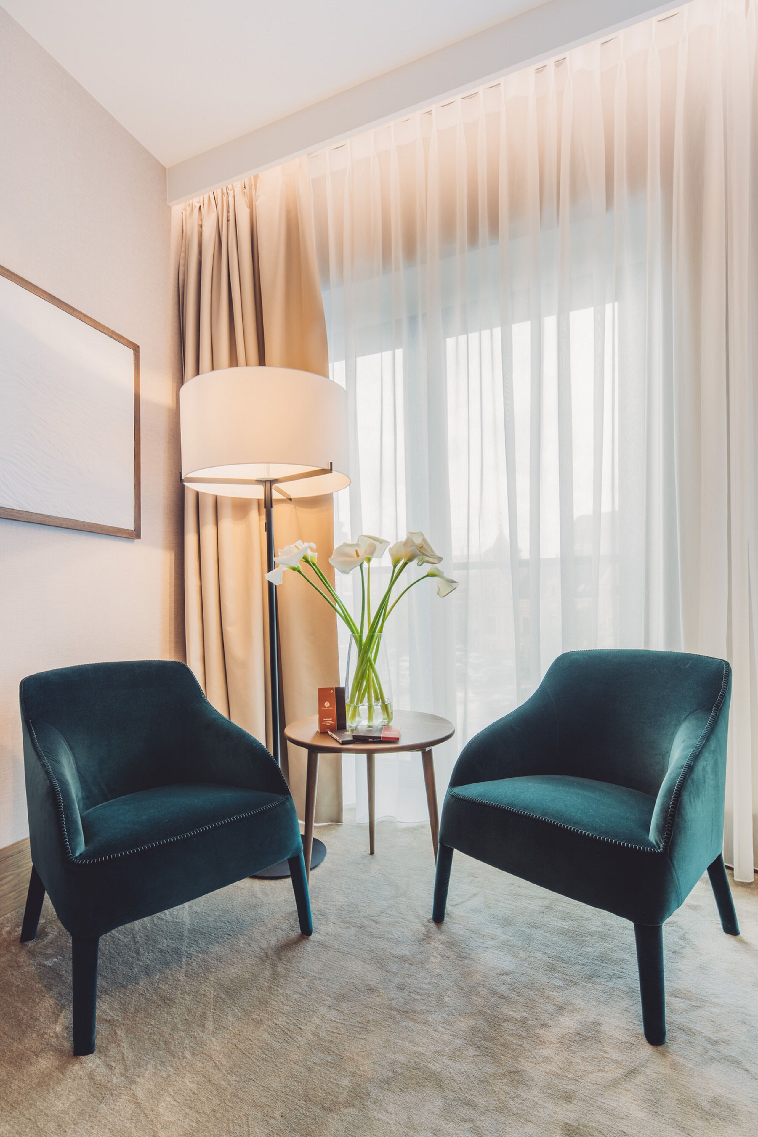 Habitación de hotel con dos sillones color turquesa, una lámpara de pie, un jarrón con flores blancas y un amplio ventanal con cortinas traslúcidas.