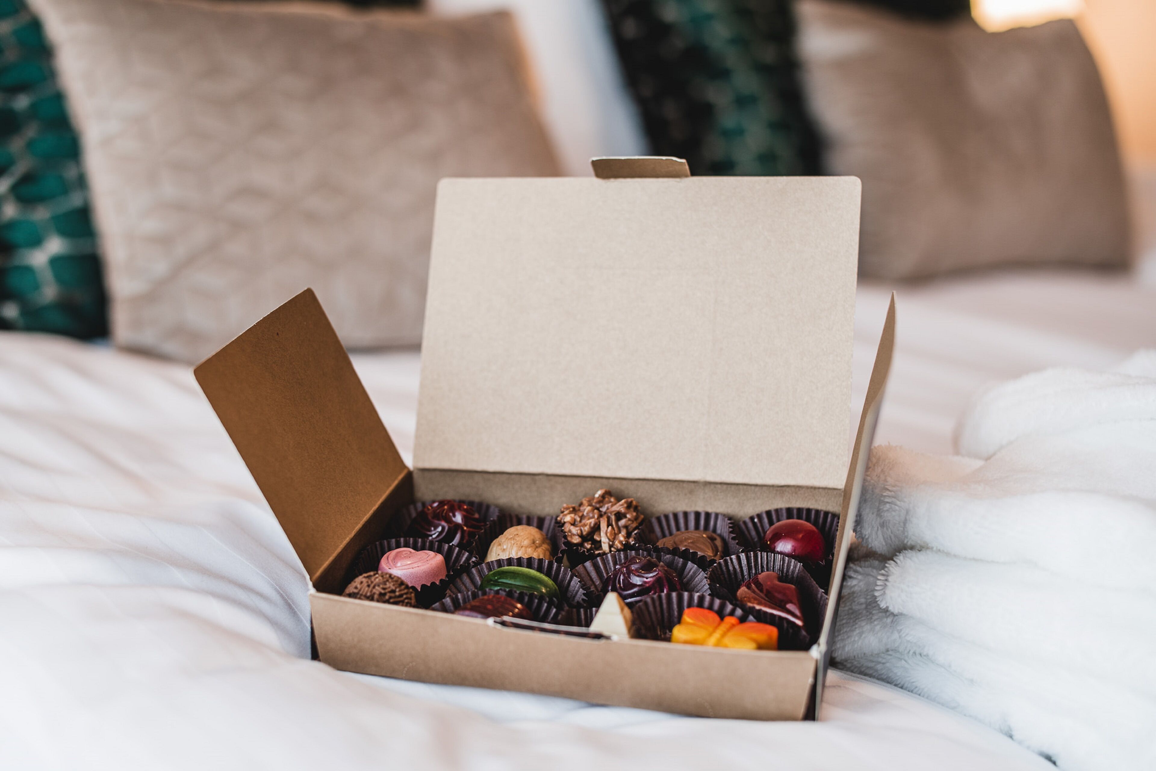A box of assorted chocolates on a bed with pillows in the background.