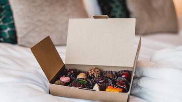 A box of assorted chocolates on a bed with pillows in the background.