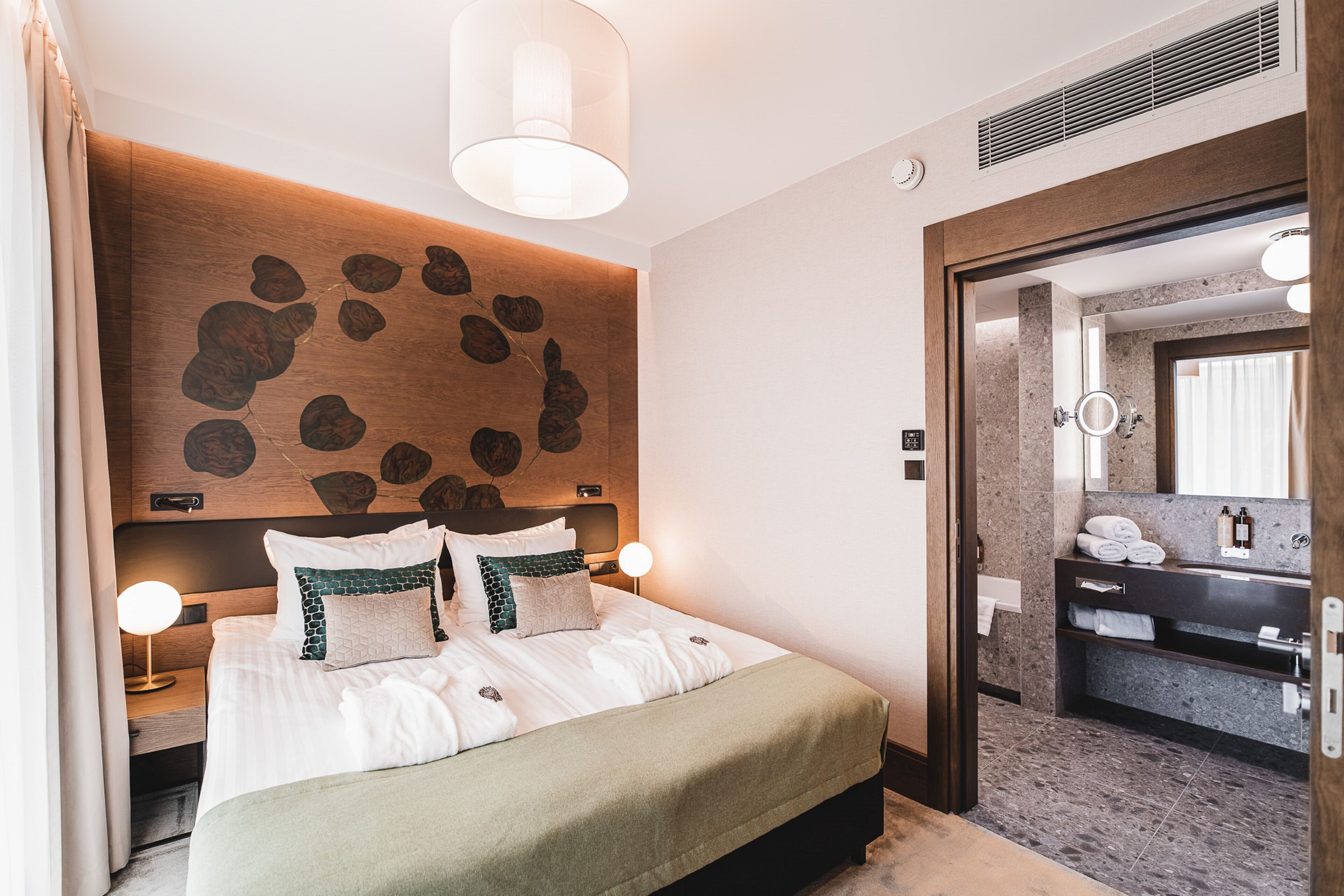 Una habitación de hotel moderna con una cama grande, un cabecero de madera con diseño de hojas, una mesita de noche con lámpara y un baño visible a través de una puerta abierta.