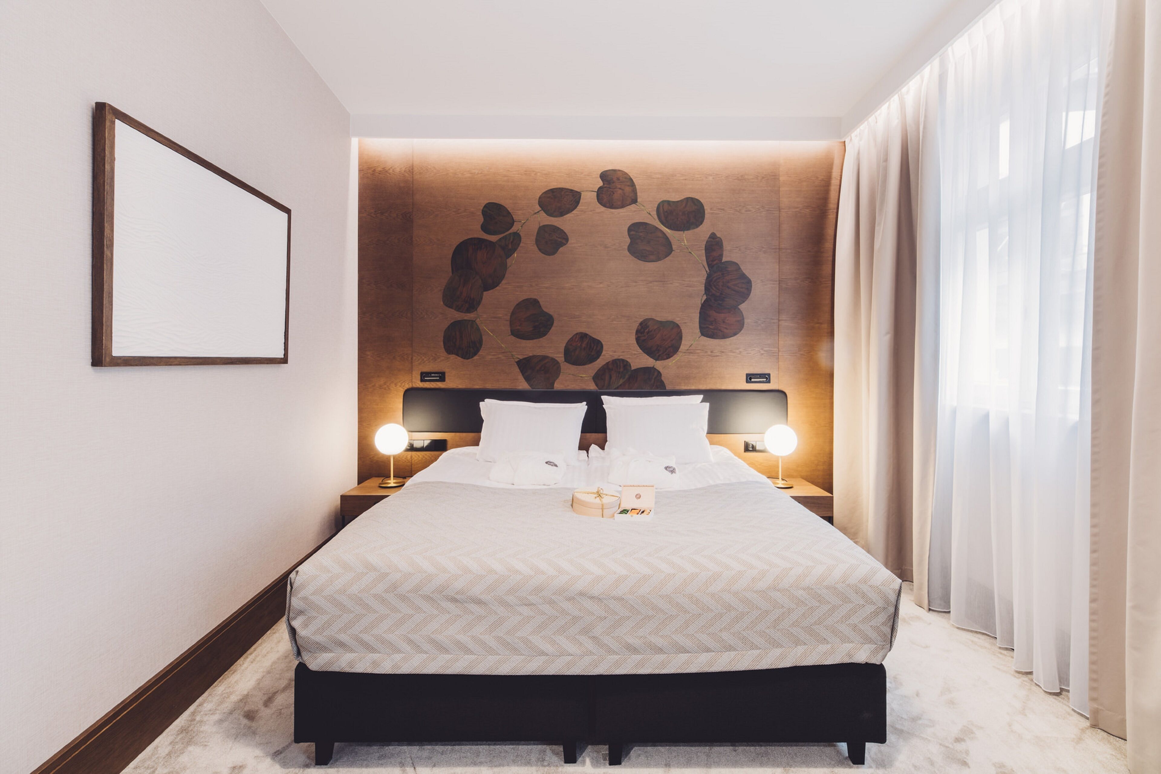Una habitación de hotel impecable con una cama grande, dos mesitas de noche con lámparas, una obra de arte en la pared y cortinas translúcidas.