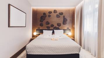 Una habitación de hotel impecable con una cama grande, dos mesitas de noche con lámparas, una obra de arte en la pared y cortinas translúcidas.