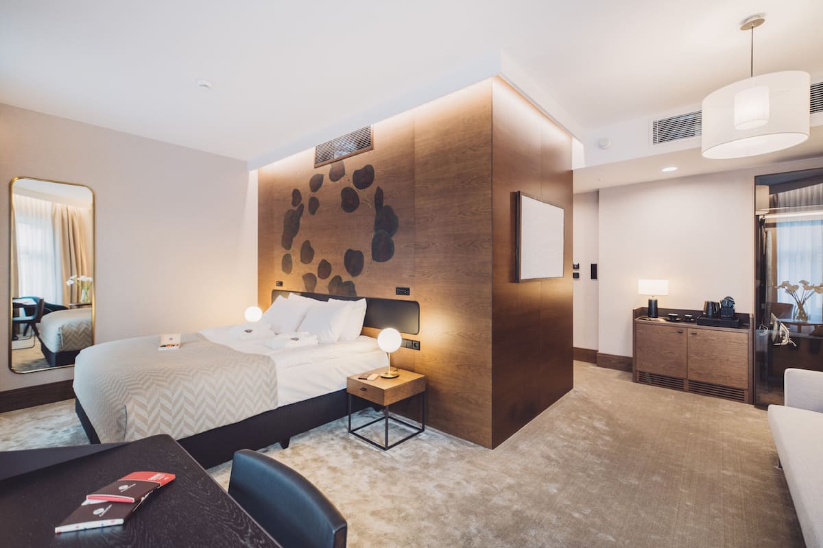 A modern hotel room with a large bed, a bedside table with a lamp, a small desk with a chair, a wooden accent wall, and a large mirror.