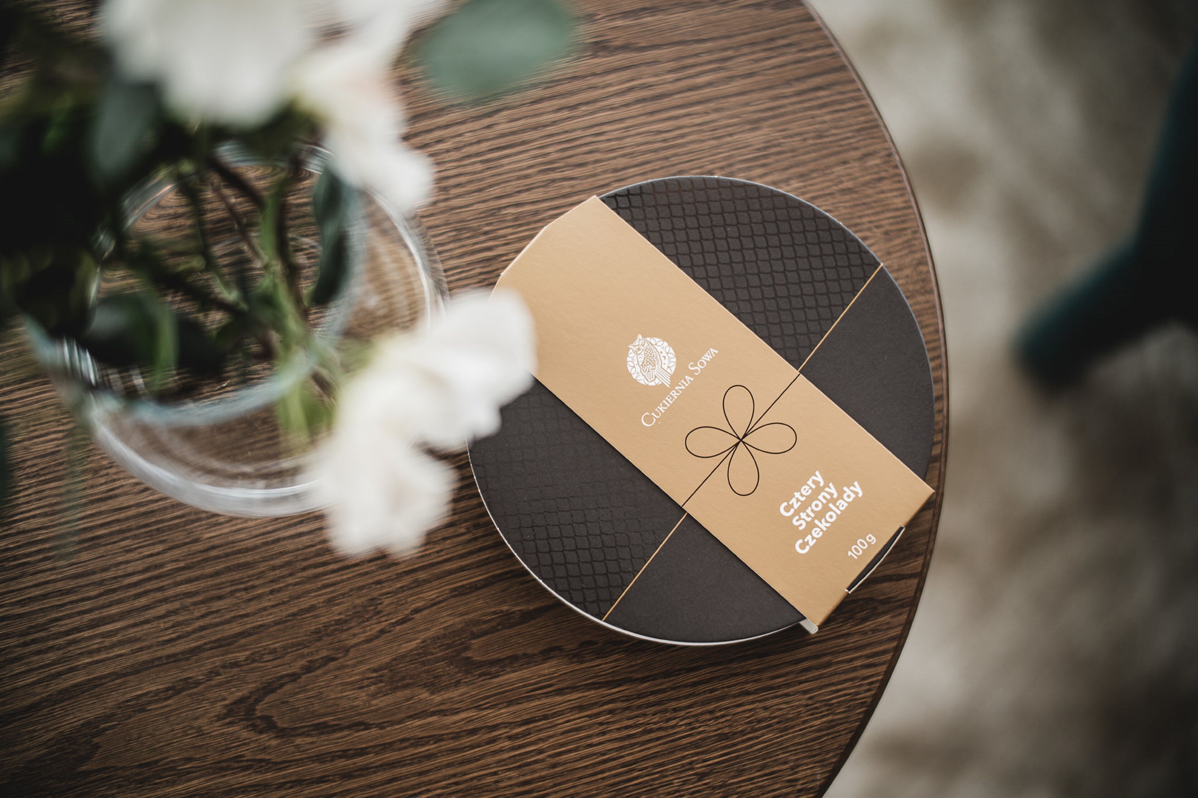 Una mesa redonda de madera con una caja etiquetada “Czech story Czełoklady” y un jarrón con flores blancas.