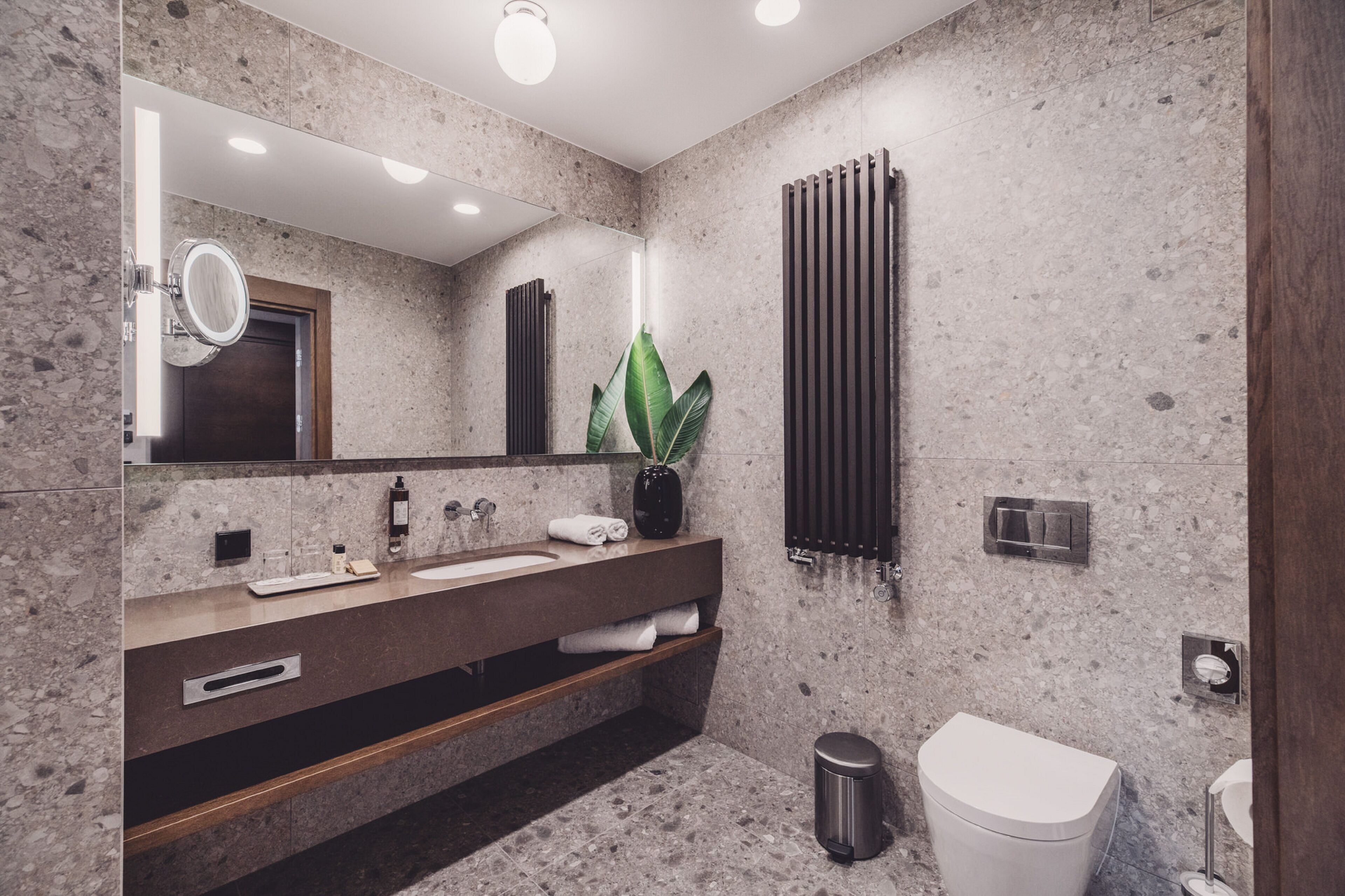 A modern bathroom with a granite countertop, a large mirror, a potted plant, a towel rack, and a toilet.