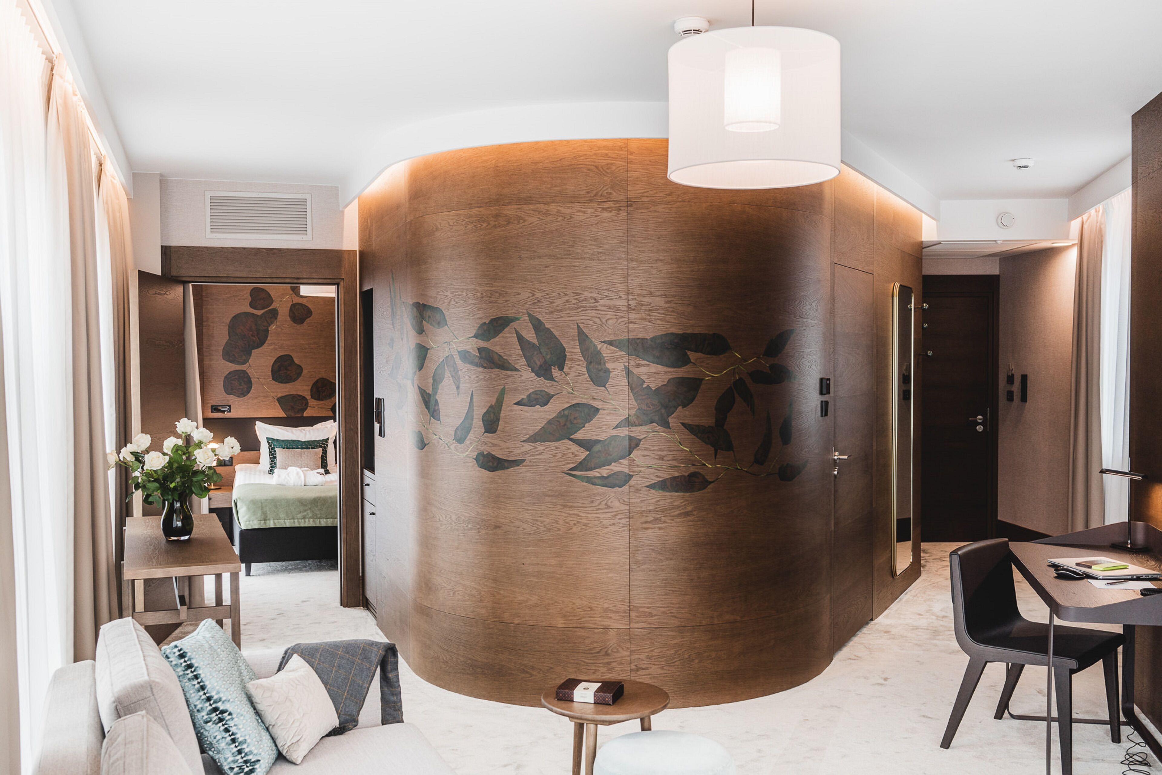 Une chambre d’hôtel moderne avec un grand mur décoratif en bois, un coin repas avec une table et des chaises, un canapé avec des coussins et une petite table d’appoint agrémentée d’un vase de fleurs.