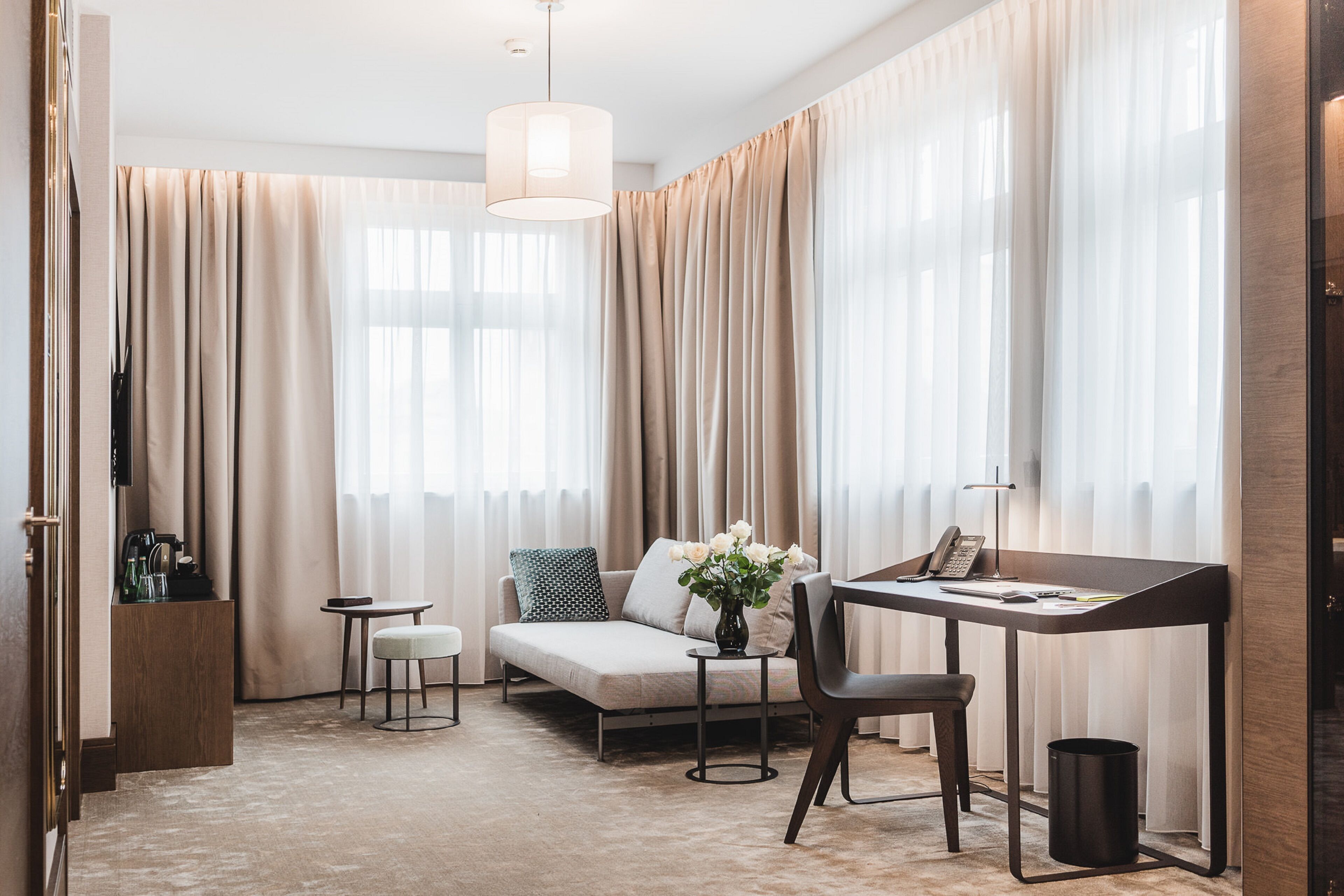 A modern hotel room with a sofa, a small table with a vase of flowers, a desk with a chair, and a lamp.