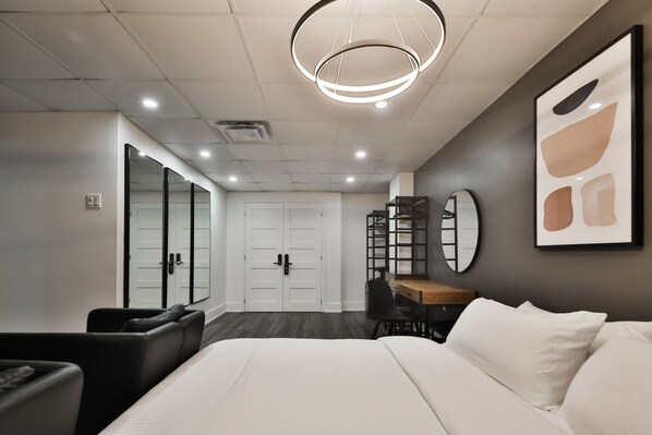 A modern hotel room with a large bed, a desk with a chair, a wardrobe, and a mirror on the wall.