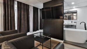 Une chambre d’hôtel moderne avec un canapé, une table en verre, un grand miroir et une salle de bain équipée d’un lavabo et d’un miroir.