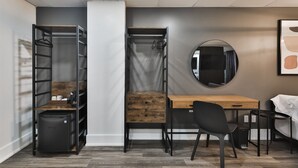 Une pièce de taille modeste, comprenant un bureau en bois, une chaise noire, une armoire avec un miroir et une petite poubelle.