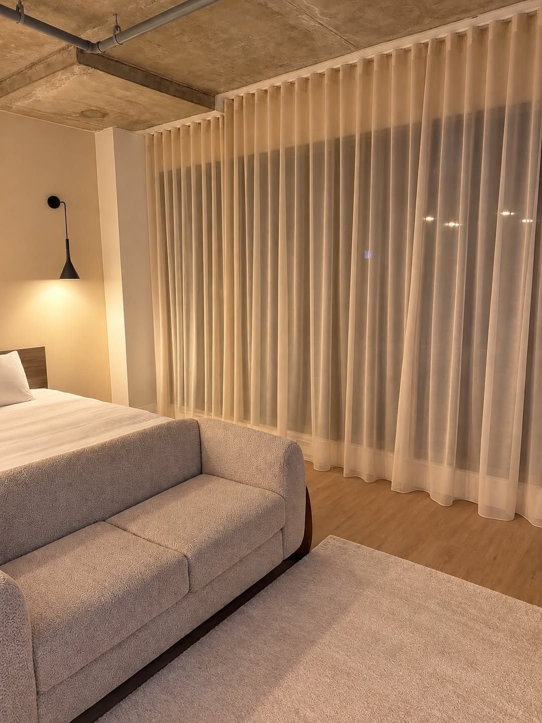 Une chambre à coucher moderne avec un lit, un canapé, une lampe et des rideaux légers.